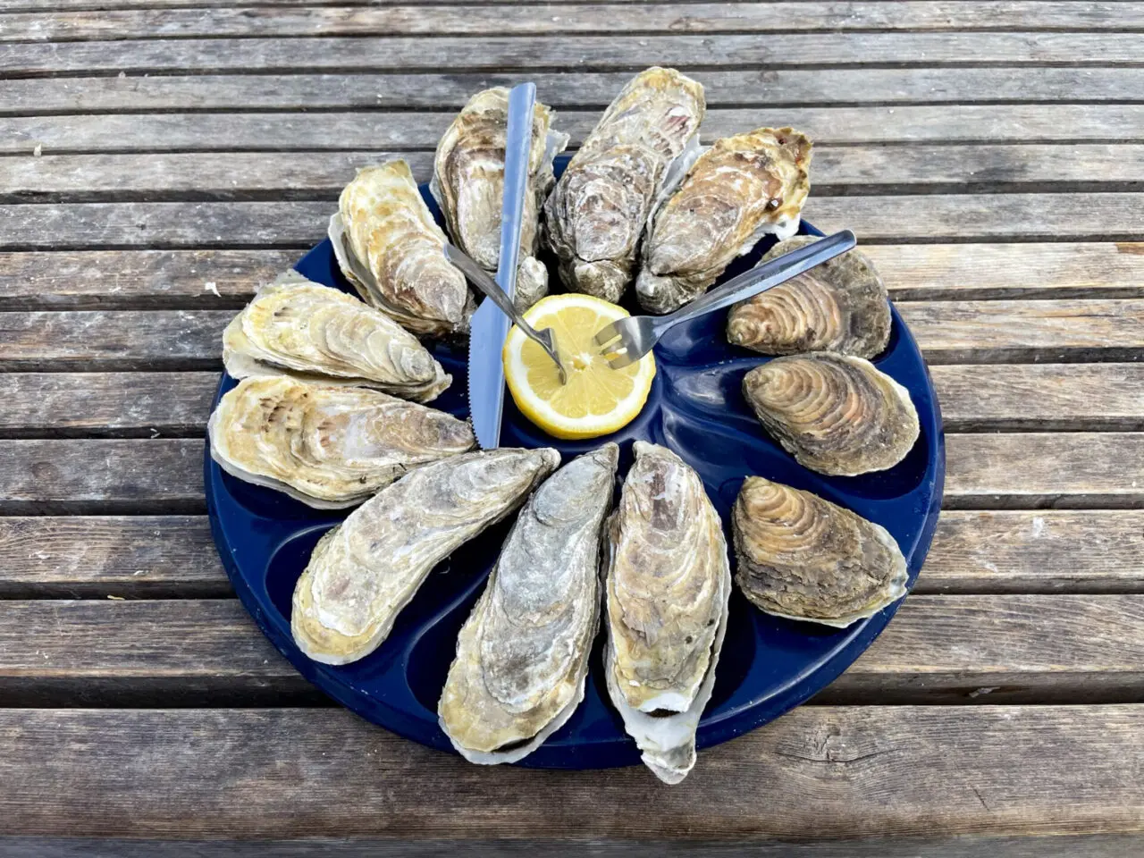 Circular platter of fresh Cancale oysters arranged around lemon wedge with oyster knives on wooden deck, showcasing premium Brittany seafood