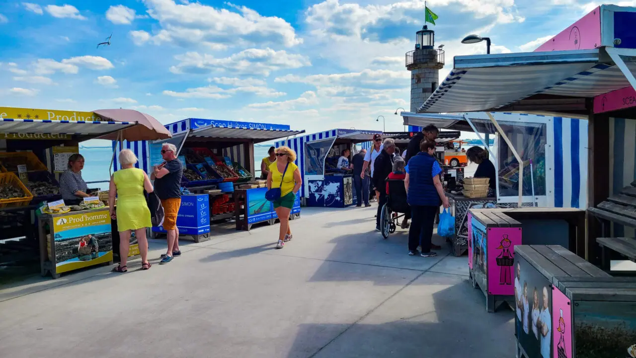 Colorful seafood market stalls at Marché aux Huîtres de Cancale with lighthouse backdrop, tourists browsing fresh oyster vendors along Brittany coast